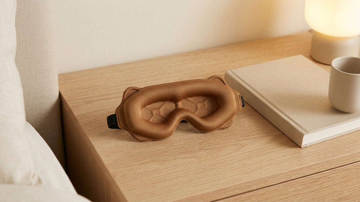 Caramel contour sleep mask with molded eye cups on an oak nightstand beside a book